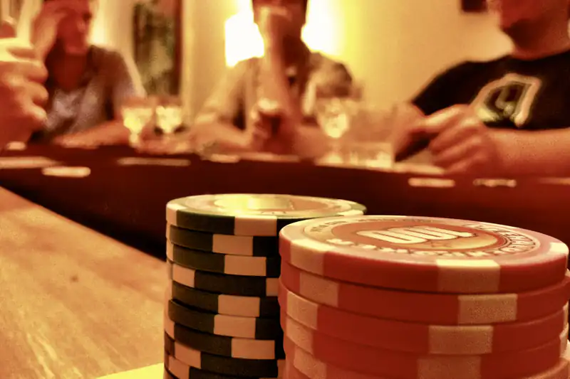 Stacks of casino chips on a table with friends playing cards in warm light